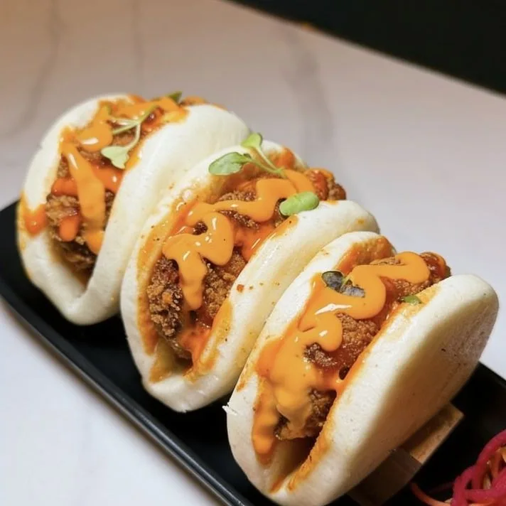 Three butter chicken baos on a black tray, topped with orange sauce and microgreens.